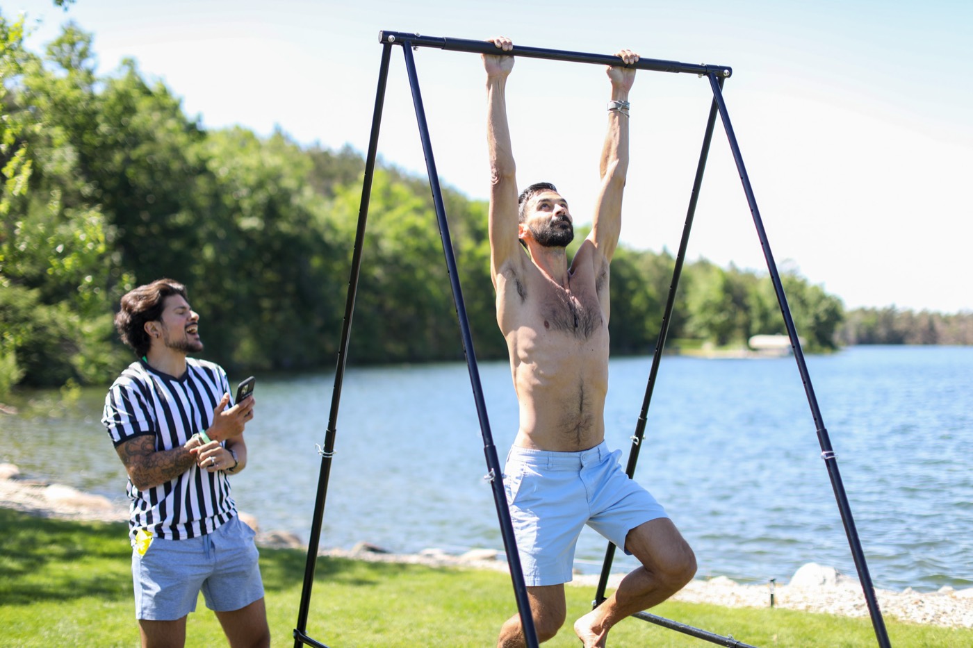 Shirtless competitor mid-pull-up at the 2026 Vegan Olympics as a referee times the reps, lake behind